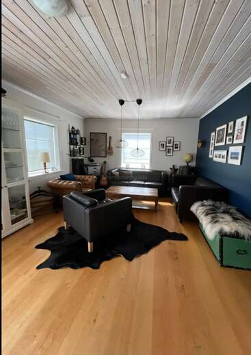 A cozy living room with wooden floors, leather chairs, a black rug, coffee table, wall art, large windows, and a blue accent wall with framed photos.