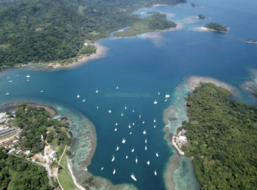 This aerial image shows a lush green coastline with turquoise waters, numerous anchored sailboats, coral reefs, small buildings, and winding waterways surrounded by tropical forest.