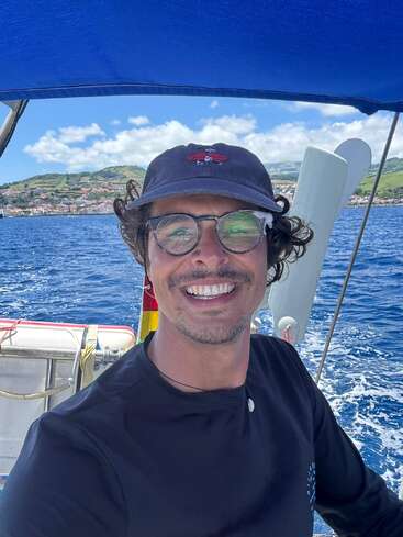 A smiling man wearing glasses and a cap takes a selfie on a boat, with vibrant blue water and a scenic green coastline in the background.
