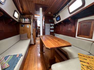 This cozy boat interior features wooden walls, a polished table, cushioned benches, small windows, bookshelves, plates on racks, and a nautical, homey atmosphere throughout.