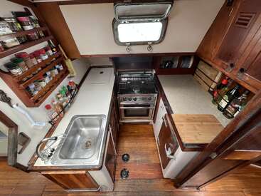 This cozy boat galley features a compact stove, stainless sink, extensive spice rack, organized bottles, wooden cabinets, porthole window, and efficient use of space.
