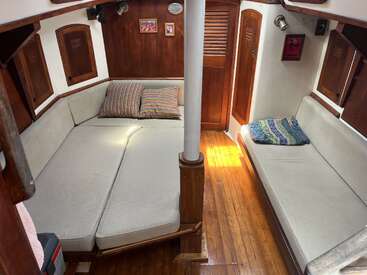 This cozy boat cabin features cushioned seating, a central support pole, polished wood paneling and floors, colorful pillows, small wall art, and ambient natural light.