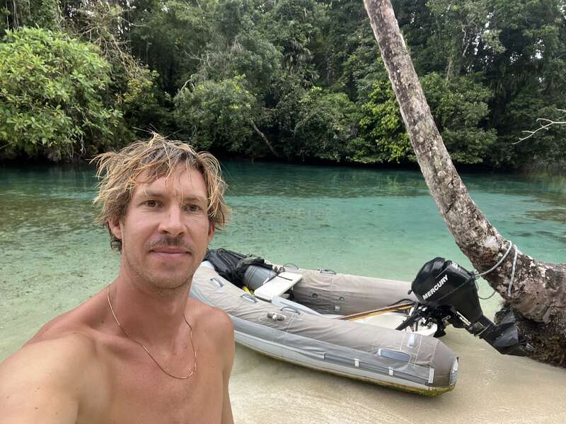 A shirtless man takes a selfie by a clear turquoise river. An inflatable boat with an outboard motor is tied to a leaning tree nearby. Lush jungle surrounds.