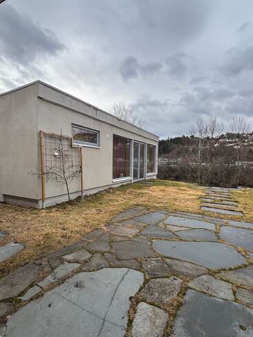 Ein modernes, minimalistisches Haus mit großen Glasfenstern steht auf einem unebenen Rasen. Ein Steinweg führt zum Eingang. Im Hintergrund sind ein bewölkter Himmel und kahle Bäume zu sehen.