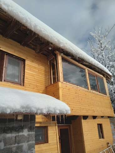 Une cabane en bois avec des toits enneigés et des stalactites qui pendent. Les fenêtres en verre reflètent le ciel hivernal. Des pins et des nuages sont visibles dans l'arrière-plan froid et serein.