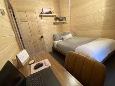 Cette chambre confortable en bois dispose d'un lit avec une literie grise, d'un bureau en bois avec un ordinateur portable et d'étagères murales où sont disposées des photos, des objets décoratifs et une plante.