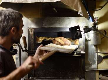 Un boulanger utilise une pelle en bois pour retirer des pains fraîchement cuits d'un four chaud, éclairé par une lampe de travail dans une cuisine rustique.