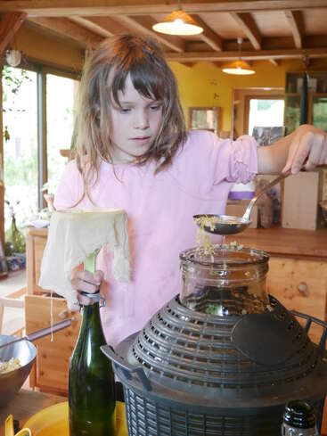Une jeune fille vêtue d'une chemise rose verse délicatement un mélange dans un grand bocal en verre, à l'aide d'une louche, tout en tenant une bouteille recouverte d'un tissu. À l'intérieur, décor rustique.