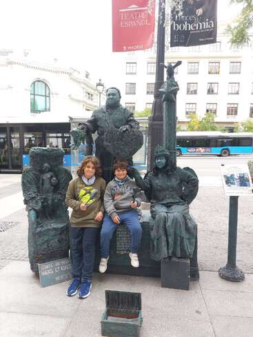 Zwei Kinder posieren mit als Statuen verkleideten Straßenkünstlern in einer städtischen Umgebung. Im Hintergrund sind ein Bus, Banner und Gebäude zu sehen, die eine lebendige Stadtatmosphäre schaffen.