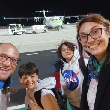 Une famille souriante de quatre personnes prend un selfie sur le tarmac d'un aéroport, la nuit, avec un avion vert et blanc visible à l'arrière-plan. L'excitation est palpable.