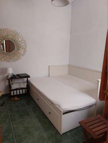 Dieses gemütliche Schlafzimmer verfügt über ein weißes Daybed mit Schubladen, einen runden Spiegel mit Blumenmuster, einen Beistelltisch mit einer Lampe, einen grün gefliesten Boden und Hausschuhe.