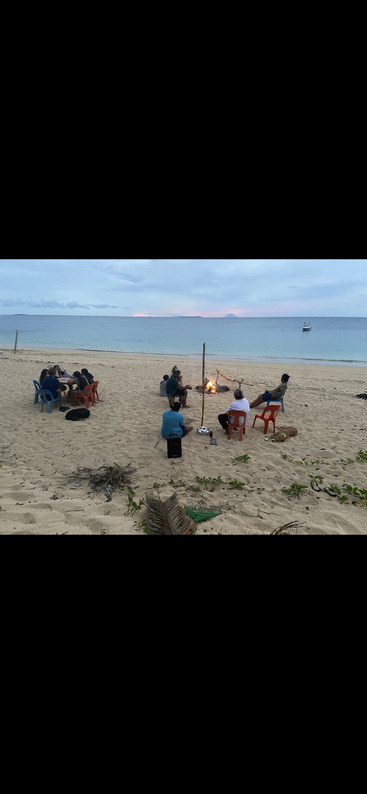 As pessoas estão sentadas em cadeiras e bancos na praia, perto do mar, reunidas em torno de uma pequena fogueira durante o pôr do sol, curtindo uma noite relaxante e tranquila juntas.