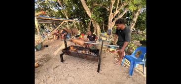 Várias pessoas estão reunidas ao ar livre, num terreno arenoso, assando um porco inteiro no espeto sobre uma fogueira, rodeadas por árvores e cadeiras de plástico coloridas. O clima é bem descontraído.