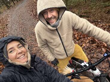 A smiling man and woman in warm jackets pose outdoors on a forest path, surrounded by autumn leaves. The man is holding a bicycle.