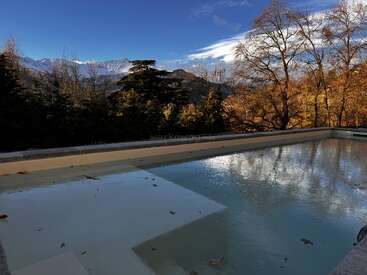 A frozen pool reflects the clear blue sky and surrounding autumn trees, with snow-capped mountains visible in the background, creating a serene and cold landscape.