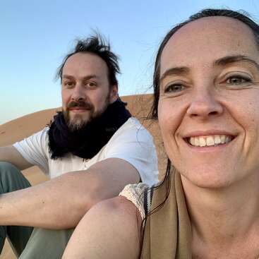 The image shows a man and woman posing for a selfie in a desert landscape, with sand dunes and a clear blue sky in the background.
