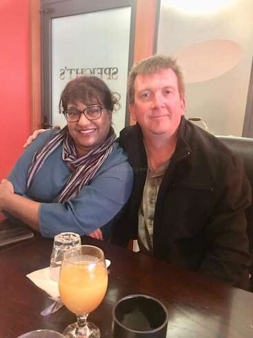 Um casal sorridente senta-se junto em uma mesa de restaurante, desfrutando da companhia um do outro. Bebidas, copos e talheres estão bem arrumados na frente deles.
