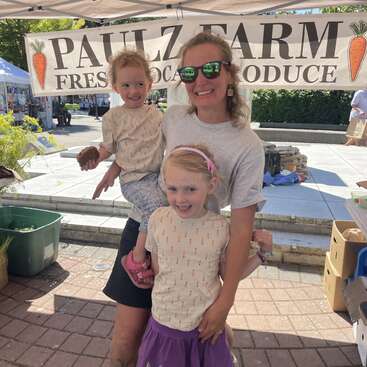 A smiling woman and two children pose together at a farmers market stand. The banner behind them reads “PAULZ FARM FRESH LOCAL PRODUCE” with carrot illustrations.