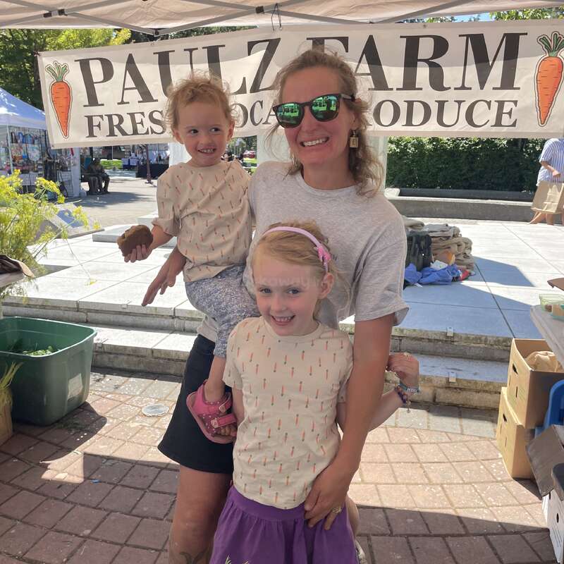 A smiling woman and two children pose together at a farmers market stand. The banner behind them reads “PAULZ FARM FRESH LOCAL PRODUCE” with carrot illustrations.