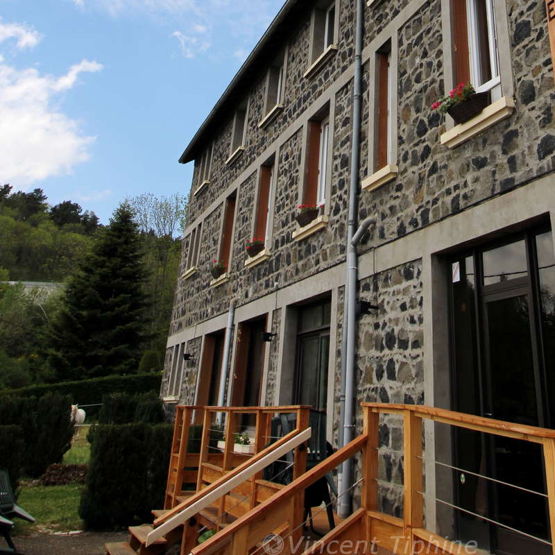 The image depicts a stone building with a wooden staircase and railing, featuring windows and a door, set against a backdrop of trees and a blue sky with clouds.