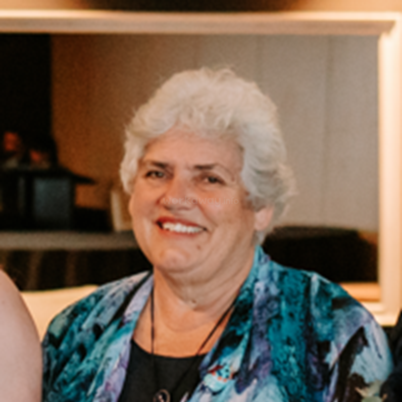 The image shows an older woman with short, curly white hair smiling warmly. She wears a colorful blue and purple top, necklace, and stands indoors, appearing happy.