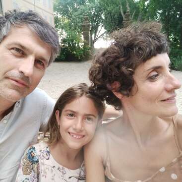 Une famille de trois personnes, composée d'un homme, d'une femme et d'une petite fille souriante, prend un selfie en plein air par une journée ensoleillée. On aperçoit des arbres et un immeuble derrière eux.
