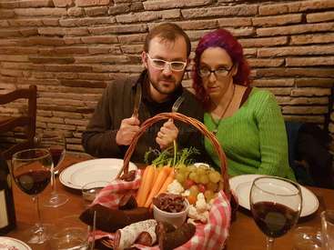 Das Bild zeigt ein Paar, das mit einem Korb voller Essen und Weingläsern an einem Tisch sitzt, vor dem Hintergrund einer Backsteinmauer.