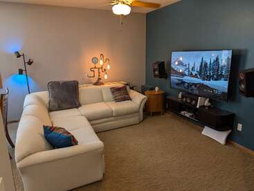 Ein gemütliches Wohnzimmer mit einem weißen Ecksofa, bunten Kissen, moderner Beleuchtung, einem an der Wand montierten Fernseher, Surround-Sound-Lautsprechern und einer blauen Akzentwand mit Wintermotiv.