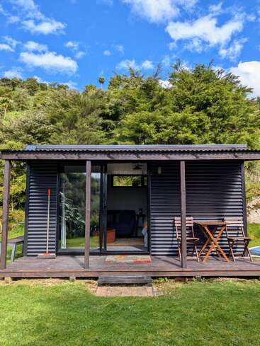 A cozy, modern tiny house sits on a wooden deck, surrounded by lush greenery and blue sky. There’s outdoor seating and a broom leaning inside.
