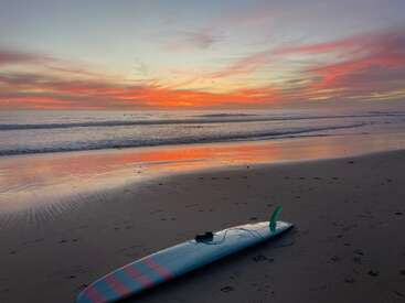 Uma prancha de surf repousa sobre a areia molhada da praia. O céu vibrante do pôr do sol reflete tons de laranja, rosa e roxo sobre o oceano calmo e a costa.