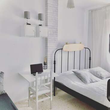 Essa imagem mostra um quarto minimalista com uma cama de metal preta, cabeceira de skate, roupa de cama branca, uma pequena mesa, cadeira, laptop, garrafa de champanhe e luzes na parede.