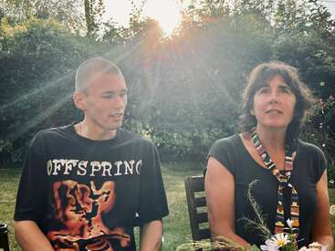 Ein junger Mann und eine junge Frau sitzen draußen an einem Tisch, mit der Sonne im Rücken. Sie sehen total entspannt aus und genießen einfach ihre Zeit zusammen in einem wunderschönen Garten.
