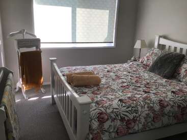 Un acogedor dormitorio con una colcha de flores, almohadas, toallas dobladas, una lámpara, una ventana y un somier blanco. La luz se filtra suavemente a través de las persianas.