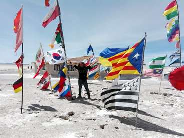 Das Bild zeigt einen Mann, der inmitten einer Sammlung internationaler Flaggen auf einem sandigen Gelände steht, während im Hintergrund ein Gebäude und Menschen unter einem klaren Himmel zu sehen sind.