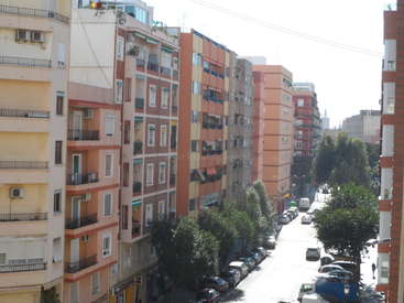 Das Bild zeigt eine von hohen Gebäuden gesäumte Stadtstraße mit entlang der Straße geparkten Autos und Bäumen auf dem Gehweg auf der rechten Seite.