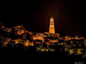 Das Bild zeigt eine kleine Stadt bei Nacht mit einem markanten Turm und zahlreichen Gebäuden, die von Bäumen und Pflanzen umgeben sind.