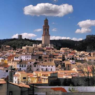 Das Bild zeigt eine kleine Stadt mit einem markanten Kirchturm, umgeben von Gebäuden und Bäumen, vor einem blauen Himmel mit weißen Wolken.