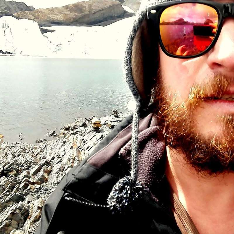 Das Bild zeigt einen Mann mit Bart und Sonnenbrille, der vor einem ruhigen Gewässer steht, umgeben von Felsen und schneebedeckten Bergen im Hintergrund.