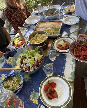 Un grupo se sienta alrededor de una mesa con varios platos: ensalada, cuscús, lasaña, tomates frescos, agua y lentejas, disfrutando juntos de una soleada comida al aire libre.