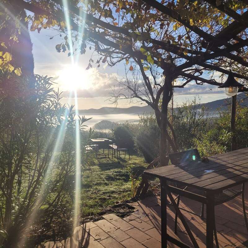 La imagen representa un sereno patio al aire libre con una mesa y sillas de madera, rodeado de frondosos árboles y arbustos, con vistas a un pintoresco paisaje con montañas y un cielo nublado.