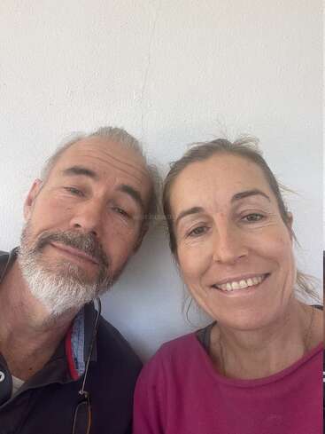 Um homem de barba grisalha e uma mulher com uma blusa rosa estão sorrindo juntos, posando bem próximos um do outro em frente a uma parede branca lisa.