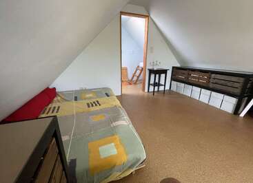 This cozy attic bedroom features slanted white walls, a single bed with a colorful duvet, simple furniture, wooden crates for storage, and a small adjoining room.