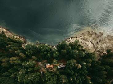 The image depicts an aerial view of a serene lake, surrounded by lush trees and a small house or cabin, with a dirt road leading to it.