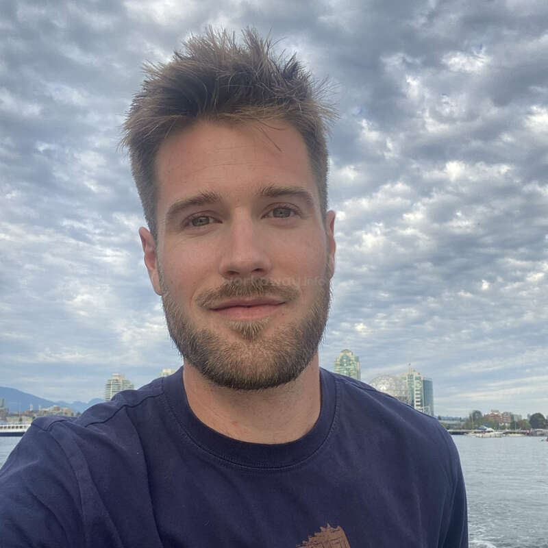 The image depicts a man with short hair and a beard, wearing a navy blue shirt, standing in front of a body of water and a city skyline under a cloudy sky.