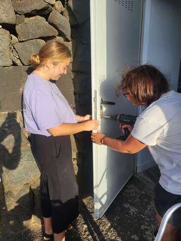 Two women are working together to repair or install a lock on a white metal door. One uses a drill while the other holds the door steady.