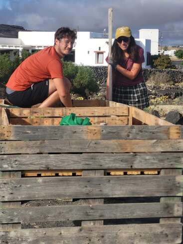 Two people are working together outdoors, possibly on a gardening or building project, using wooden pallets. Both are smiling, enjoying a sunny day with cloudy skies.