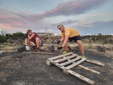 Two people are outdoors building a wooden structure using pallets and tools. Both are smiling, wearing casual clothes and hats, under a colorful sunset sky.