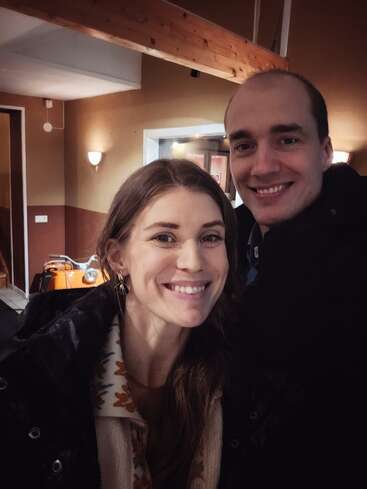 Ein lächelndes Paar posiert drinnen, warm gekleidet in Jacken. Der gemütliche Raum hat braune Wände, Holzbalken, sanfte Beleuchtung und ein orangefarbenes Fahrzeug im Hintergrund.