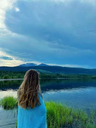 Una mujer envuelta en una manta azul está de pie junto a un sereno lago, contemplando las lejanas montañas bajo un dramático cielo azul nublado, rodeada de una naturaleza tranquila.