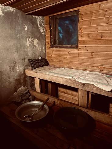 This image shows a rustic, dimly lit room with wooden walls and ceiling, a bench with a pillow, towels, and metal bowls, likely a sauna.